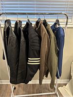 Six men's jackets hanging on white clothing rack before window blind. Various materials and colors including leather, fabric, and corduroy. Range of colors black, brown, green, blue and one jacket has white sleeve stripes.