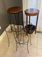 Five plant stands with one wooden top stand with metal legs, one wooden round stand, and three black metal wire stands, arranged on a tiled floor near a window.