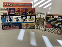 Lot showing stacked boxes: Pokemon Trainer, Kenworth truck model kit by Revell, Axis & Allies board game, Tamiya Military Miniatures, and 1991 Nascar collectible set.