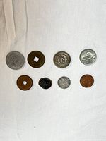 Top view of 8 foreign coins arranged on white cloth, varying metals and sizes, including copper, silver, bronze colors and square holes in two coins.