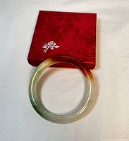 Natural multi-colored jadeite bracelet displayed next to a red display case with floral design on top. Bracelet shows gradient color tones: orange, green, and white.