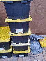 Stack of four black plastic storage totes with yellow lids, multiple lids shown beside them on ground