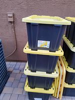 Stacked black HDX 27 gallon storage totes with yellow lids, shown outdoors on a paved surface against a textured wall.
