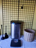 Photo showing a large automatic stainless steel kitchen garbage can with black lid, black plastic toilet brush and plunger, mechanical weight scale with black base and red dial, and a woven wicker garbage basket with natural finish placed on a white cloth against a metal grid background.