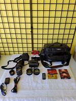 Photo of whole lot laid out on white surface against black wire grid background: includes black camera lenses, Ambico camera bag, black handheld camera, remote control, video tapes, charger, and cables.