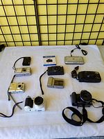 Lot of nine cameras and accessories displayed on white cloth with black grid background, showing different camera types, sizes and colors.