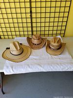 Three new cowboy hats placed on a white towel on a table against a yellow wall and black grid background. Hats are different styles with various braided and leather bands.