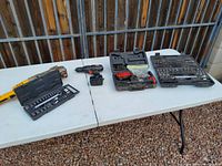 Photo of multiple tool sets on two white tables, including DeWalt cordless drill in black and yellow, Dremel rotary tool, and various socket sets, showing used condition.