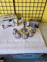 Photo showing the manual vegetable/fruit prep tool with base, crank handle, and six stainless steel drums arranged around it on a white cloth.