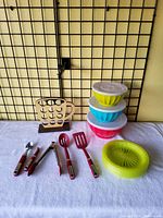 Photo showing three large plastic bowls with lids stacked in yellow, blue, and red colors, red spatula, slotted spatula, metal tongs with red handles, yellow citrus juicer bowl, and a wooden paper plate holder shaped like a coffee cup.
