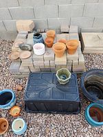 Various size planter pots and saucers arranged on gravel with pavers stacked behind. Includes terracotta, glazed ceramic in blue, white, and green hexagonal shapes. Plastic black bin and large black pot visible but not included.