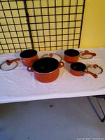 Front view of four orange pots and pans with lids on a white surface against a yellow wall with black grid background.