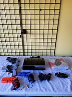 Sony PlayStation 2 slim console with multiple game controllers including two gun controllers, standard controllers, clear blue controller shell, and black multi-controller adapter all laid on white cloth.