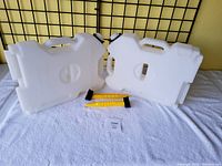 Two white Rotopax water containers laying flat with two yellow mounting brackets placed in front on a white towel against a black wire backdrop.