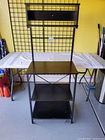 Front view of black metal shelf unit with drawer and three shelves, metal cross support visible at back.