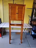 Front view of tall wooden over toilet shelf rack showing closed louvered double doors, slim legs, and flat top surface.