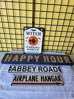 Four signs displayed: wooden Happy Hour sign at bottom with three metal signs stacked above, Witch Parking Only on top, Abbey Road below that, and Airplane Hangar at bottom.