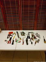 Photo showing multiple hand tools spread on a white table including pliers, screwdrivers, clamps, wrenches, sockets, and small tools.