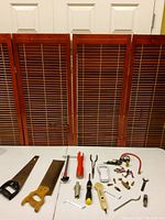 Overview of lot laid out on white surface showing hand saws, clamp tool, screwdriver, valve key, and various small tools and fittings.