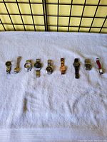 Nine various watches laid out in a horizontal line on a white towel, showing full length of each with metal and leather bands, different colors and styles.