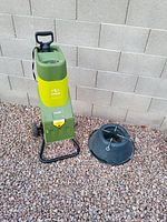 Electric wood chipper standing upright next to black Christmas tree stand, placed on gravel ground against a concrete block wall.