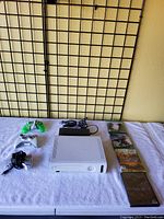 White Xbox 360 console, two wired controllers (white and green), power supply, and four Xbox 360 game cases all displayed on white cloth.