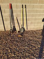 Full view of yard tools including shovel, rake, hole digger with red handles, and other hand tools against rock ground and block wall background.