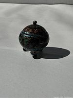 Side view of round cloisonne enamel trinket box with three feet and elephant design under natural light casting shadow.