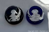 Two round glass paperweights side by side, black and dark blue, each with white engraved portraits of historical figures and decorative banners.