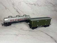 Two HO scale model train cars pictured side by side on a marble-effect background, one silver Republic tanker and one green boxcar.