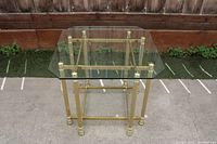 Clear top gold metal framed end table with stepped legs and crossbars, round foot details, shown outdoors against wooden fence.
