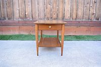Front view of mid-century nightstand on concrete with wooden fence backdrop, showing drawer with brass knob and tapered legs.