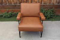 Front view of the mid century lounge chair showing brown leather cushions, padded armrests, and black metal legs with brass caps.