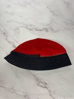 Red wool newsboy cabbie hat with shiny black bill on white marble background, front-side view.