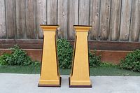 Front view of the two tapered rectangular wooden pedestal plant holders with golden-yellow finish and dark brown bases and tops, standing outdoors against a wooden fence with some greenery.