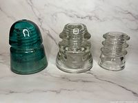 Three vintage glass electrical insulators shown side by side on a white and gray marbled background, highlighting size and color differences