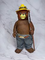 Front view of the Smokey The Bear figure wearing a yellow ranger hat labeled 'SMOKEY' and holding a shovel, dressed in gray pants with belt.