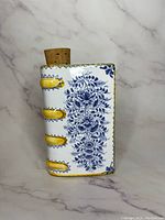 Front and side view of vintage ceramic book bottle with blue floral and yellow binding details, cork stopper on top.