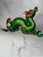 Side view of the metallic green and gold dragon figurine with red eyes and curled tail, holding a glass orb.