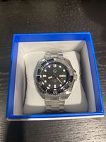 Front view of the black dial watch in a blue-lined white watch box showing stainless steel bracelet and rotating bezel.