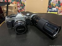 Canon AE-1 Program camera body with the 50mm f/1.8 lens mounted and lens cap on. 100-200mm zoom lens is beside it.