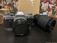 Canon AT-1 camera body with Canon lens attached and Sigma zoom lens beside it on table.