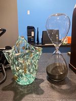 Photo shows the hourglass with black sand next to the art glass vase with multicolor design on dark surface.