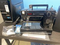 Front-facing view of black Janome sewing machine with built-in handle, showing stitch selection dial, length dial, reverse lever and removable extension table