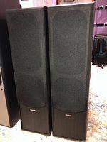 Photo of two black Quest Q660 floor-standing speakers side by side with fabric grille covers on and visible Quest logo on each.