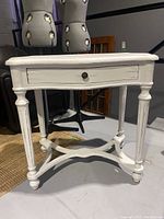 Front view of the painted white wooden side table showing turned legs, single drawer with metal knob, and sculpted apron.