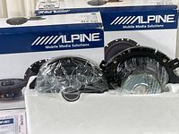 Two pairs of Alpine SPS-610 speakers with one box partially opened showing the speakers still in protective wrapping inside foam packaging and the original box with branding.