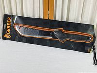 Photo of Gerber Outdoor cutting tool in unopened, taped packaging showing the full machete with saw-toothed blade and black handle.