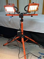 Full view of dual construction work lights mounted on orange telescoping tripod stand. Includes protective cages on lights and visible power cords connected to a control box on tripod.