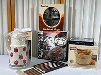 Kitchen collection items including Thermos Coffee Butler, Thermo-Serv insulated mugs, steak knife set, Rival crock pot, springform cake pan and Angel trivet in boxes and plastic wrap.